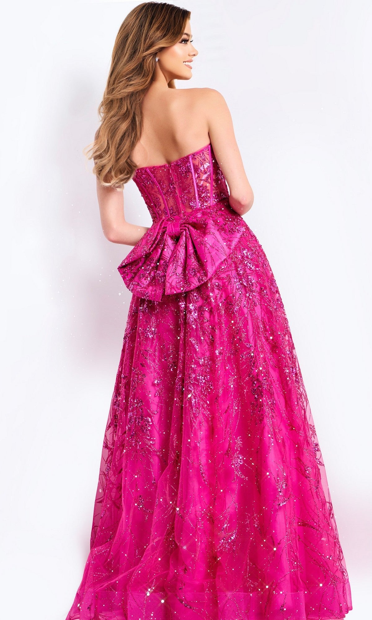 A woman with long wavy hair models the Jovani 45913 long prom dress, shown from the back. The strapless gown features a corset bodice, bright pink sequins, a large bow, and glitter details against a plain white background.